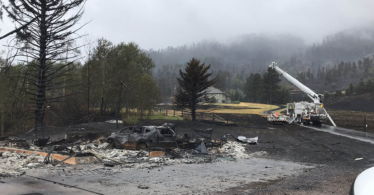 A bucket truck is seen in the background repairing a power pole after a fire. A burned car is in the foreground.