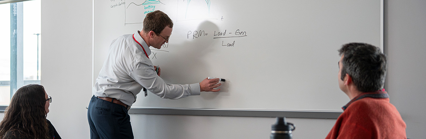 An engineer writes an equation on a whiteboard while two other engineers look on.